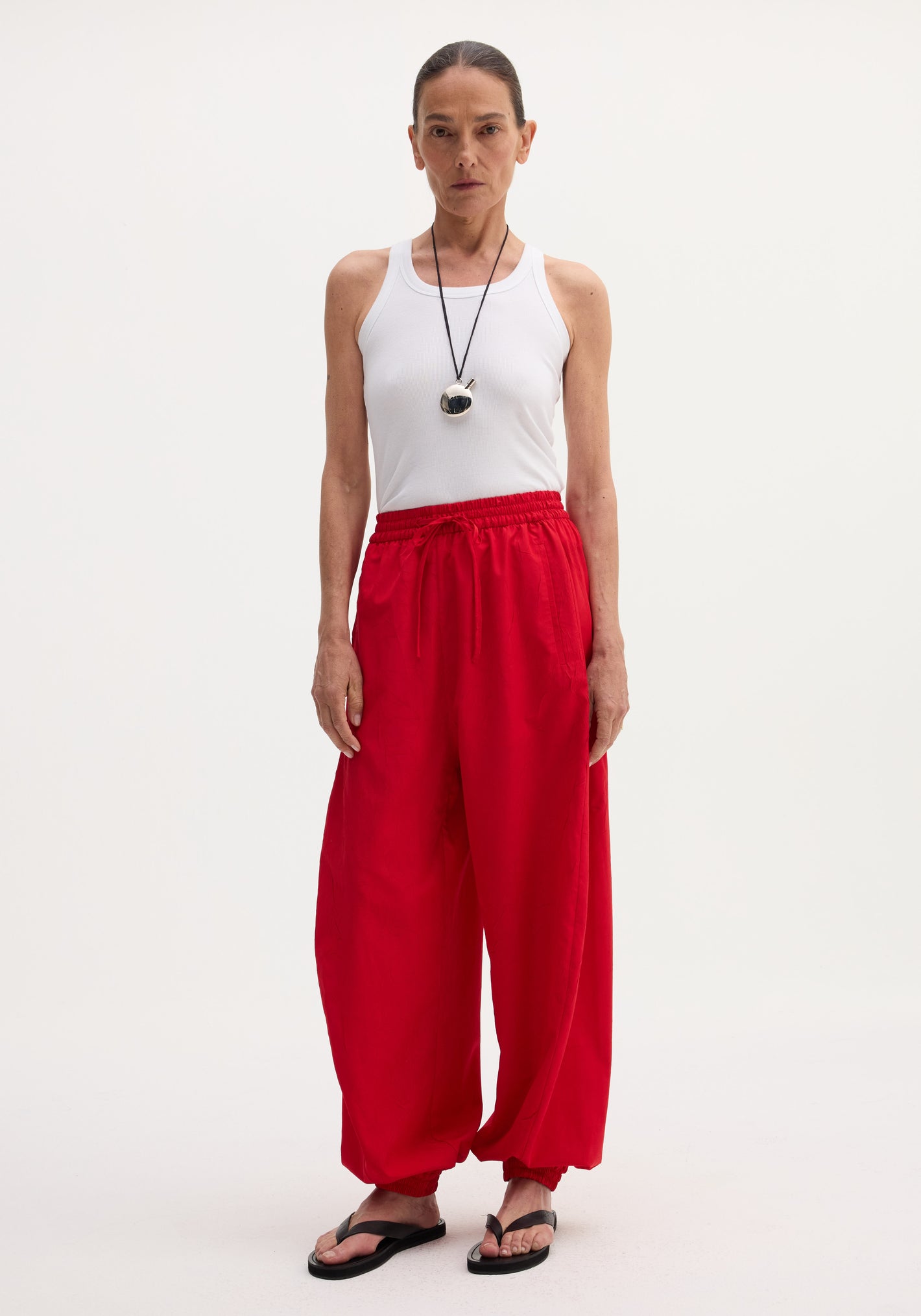 Woman wears Balloon track pants in red with elasticated waistband, drawstring, and welt pockets, paired with a white rib-tank top and black sandals.