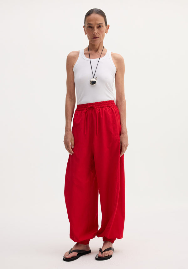 Woman wears Balloon track pants in red with elasticated waistband, drawstring, and welt pockets, paired with a white rib-tank top and black sandals.