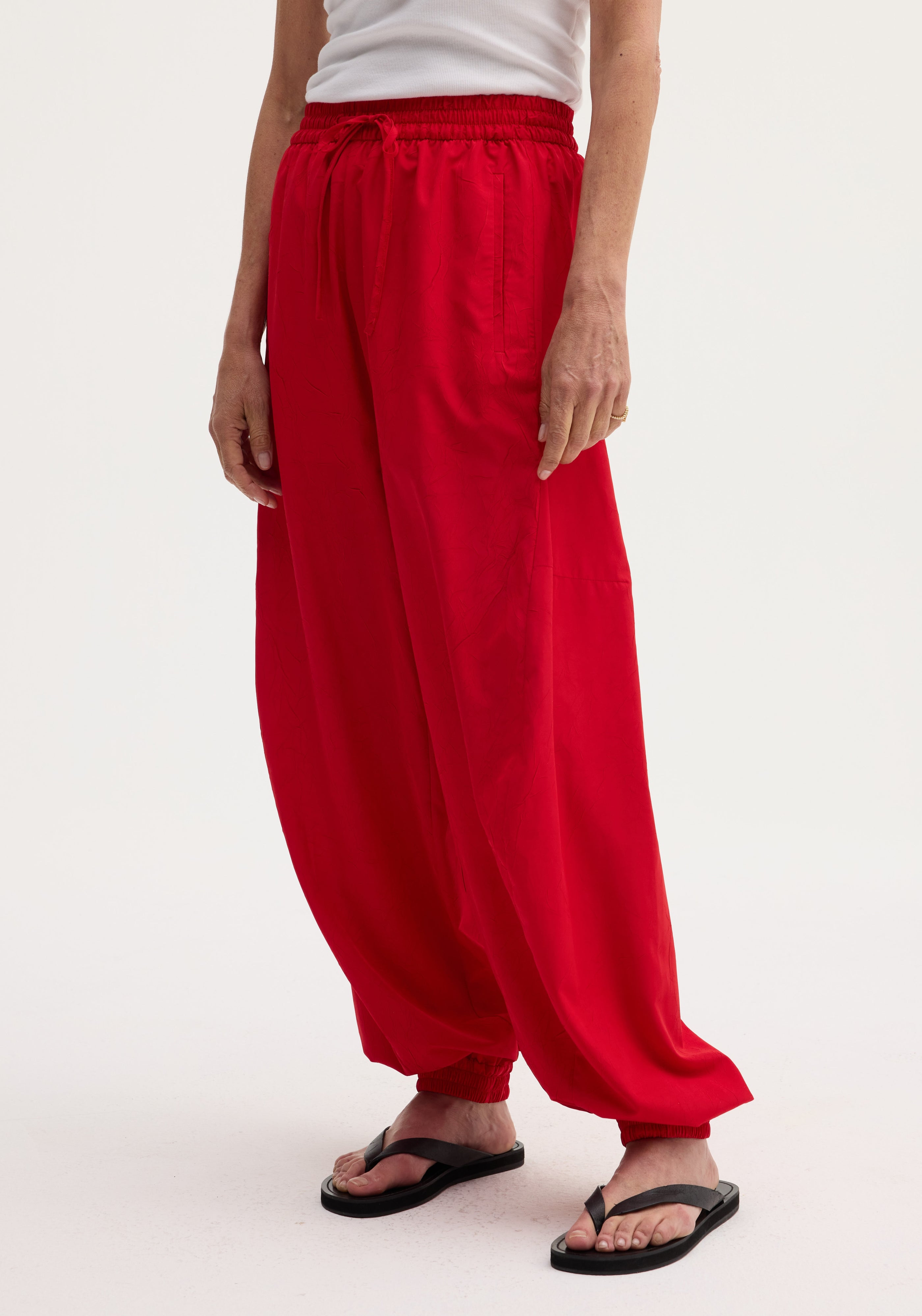 Balloon track pants in red with elasticated waistband and cuffs, featuring front welt pockets and a voluminous leg, styled with black flip-flops.