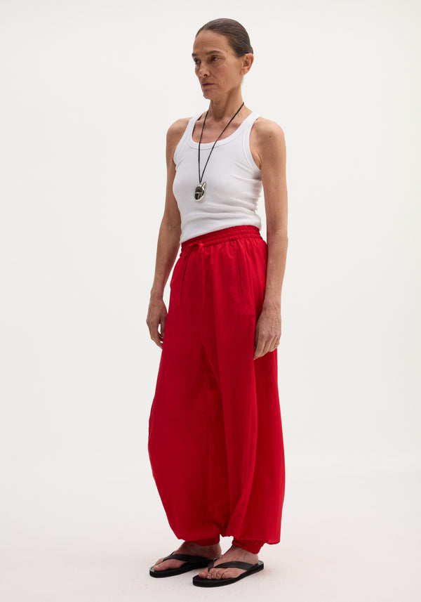 Balloon track pants in red with voluminous legs, elasticated waistband, and hems. Features front welt pockets. Model wears pants with a white tank top and flip-flops.