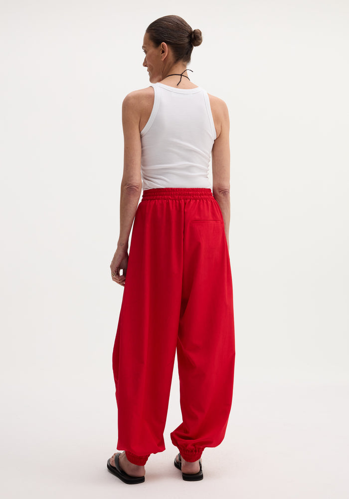 Back view of person in Balloon track pants with elastic cuffs, wearing a white tank top and black slide sandals, showcasing the pants' voluminous design.