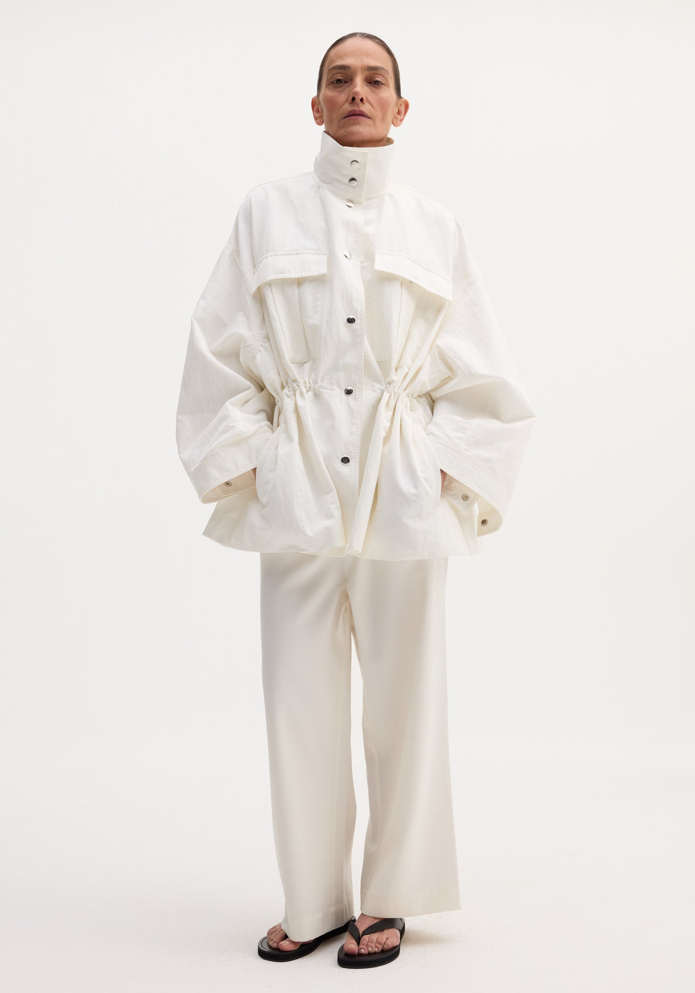 Model wearing Sculptural safari jacket in optic white, featuring a stand collar, oversized bellows pockets, and elasticated waist, paired with matching trousers and black sandals.