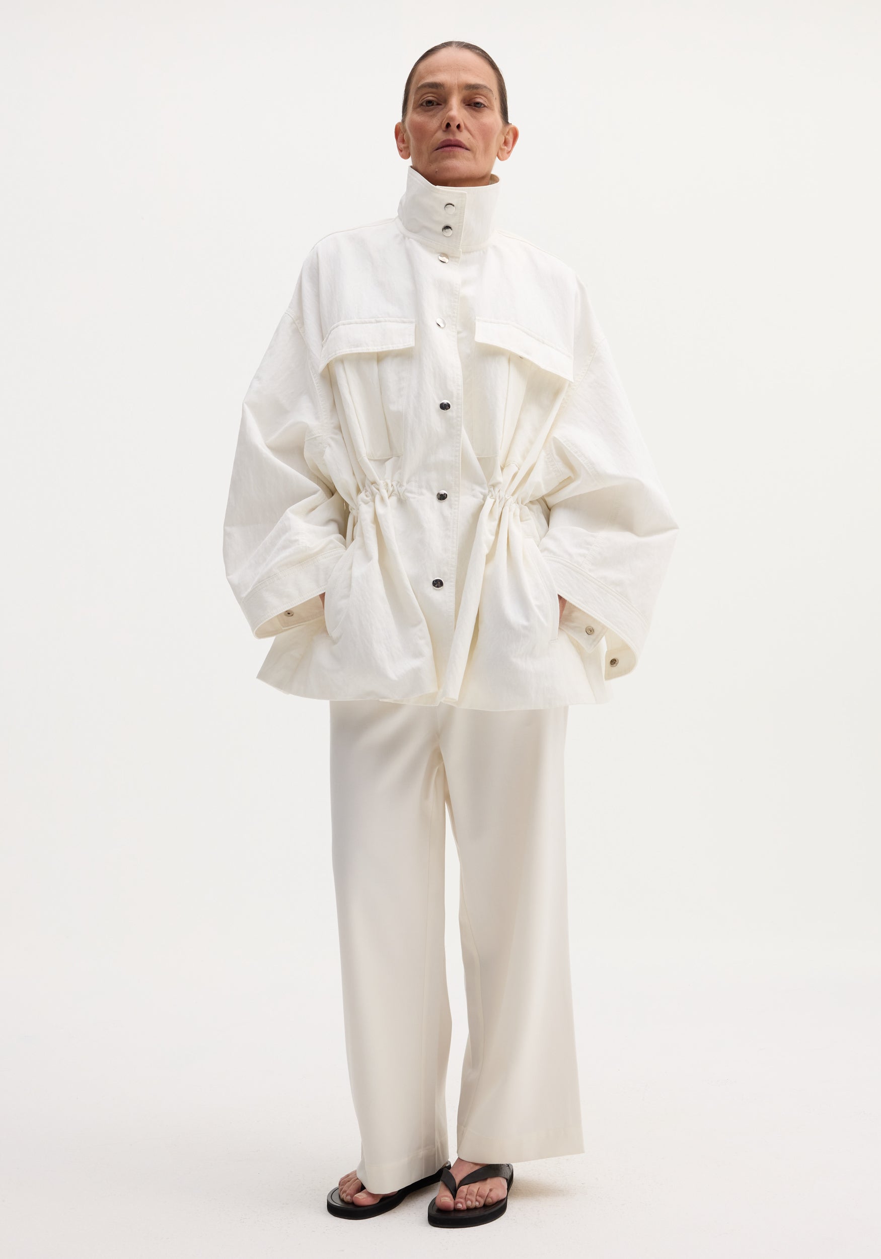 Model wearing Sculptural safari jacket in optic white, featuring a stand collar, oversized bellows pockets, and elasticated waist, paired with matching trousers and black sandals.