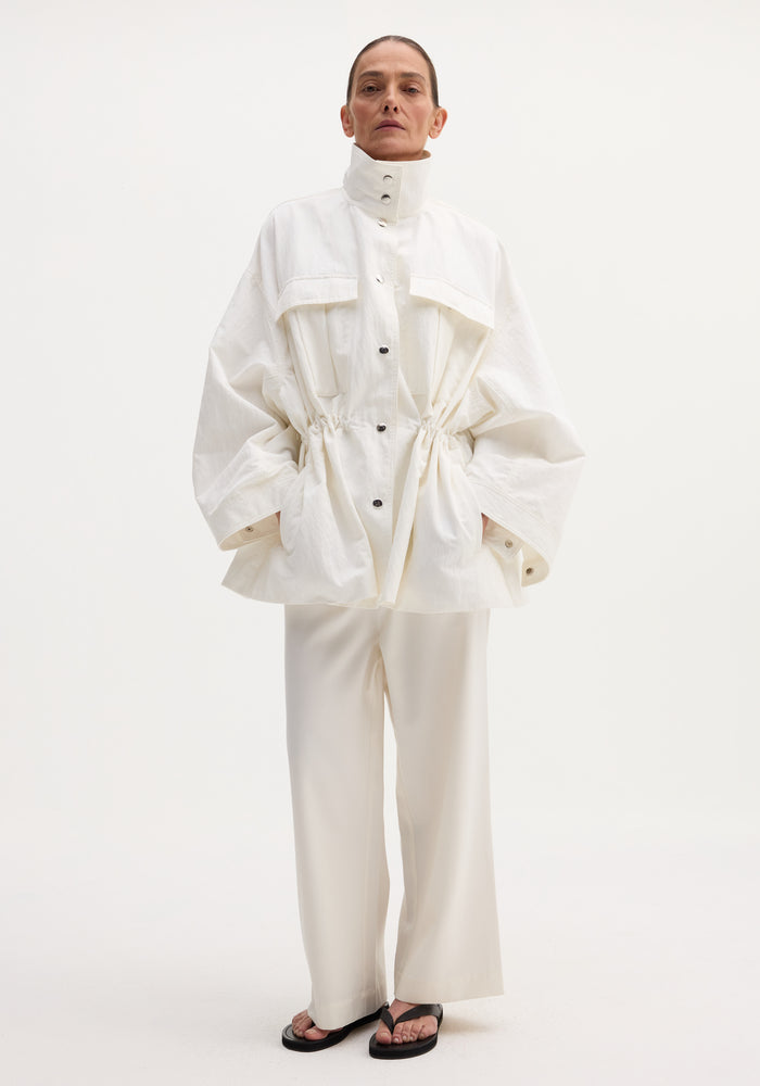 Model wearing Sculptural safari jacket in optic white, featuring a stand collar, oversized bellows pockets, and elasticated waist, paired with matching trousers and black sandals.