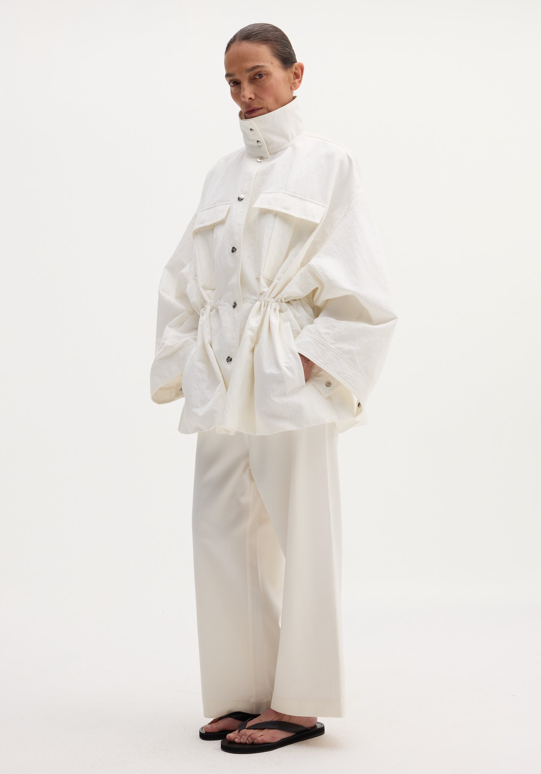 Model wearing Sculptural safari jacket with stand collar, bellows pockets, and elasticated waist, paired with matching wide-leg trousers and black thong sandals.