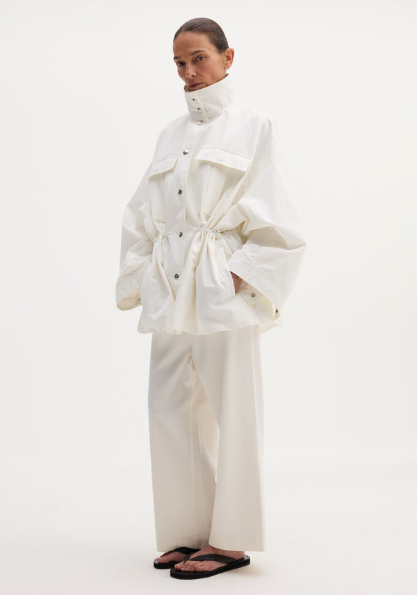 Model wearing Sculptural safari jacket with stand collar, bellows pockets, and elasticated waist, paired with matching wide-leg trousers and black thong sandals.