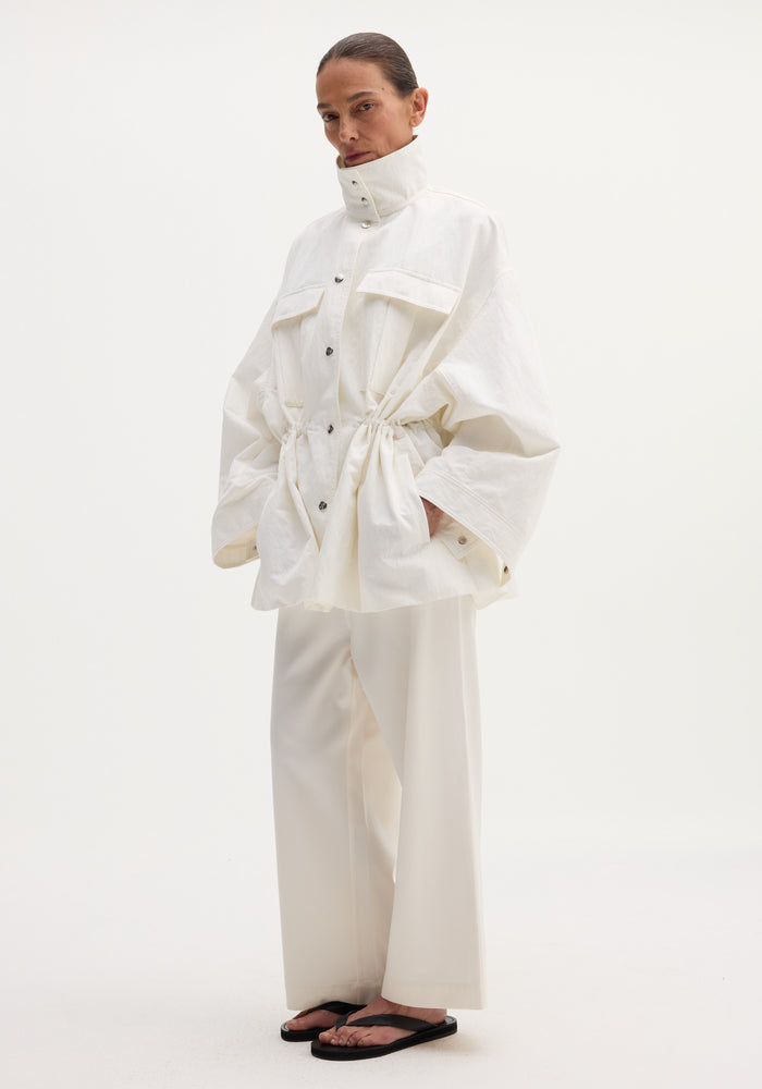 Model wearing Sculptural safari jacket with stand collar, bellows pockets, and elasticated waist, paired with matching wide-leg trousers and black thong sandals.