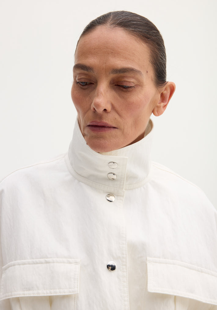 Woman in a Sculptural Safari Jacket | Optic White with stand collar and shiny snap buttons, looking contemplative.