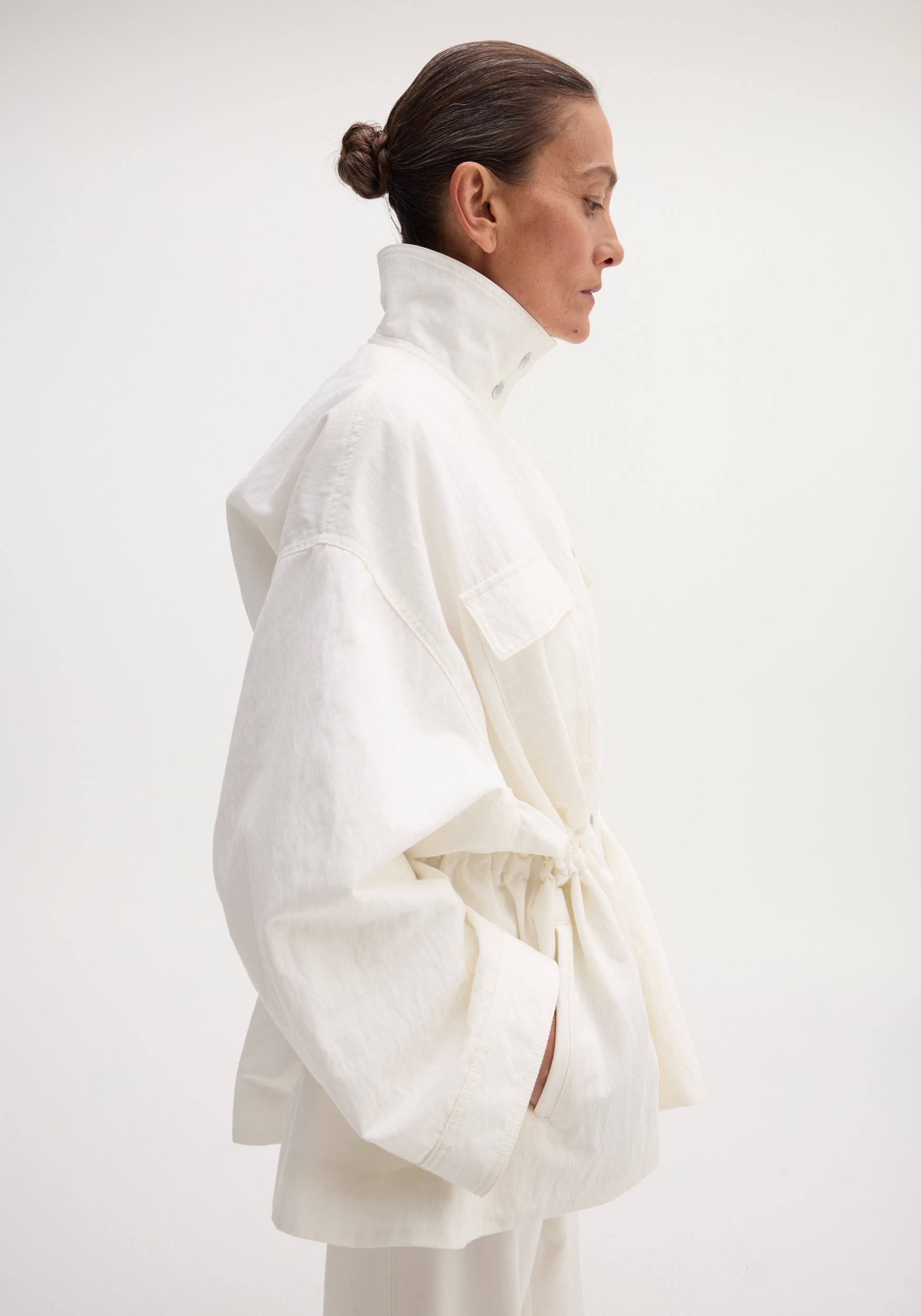 Profile view of a model wearing the Sculptural Safari Jacket in optic white, featuring a high stand collar, oversized pockets, and a cinched waist.