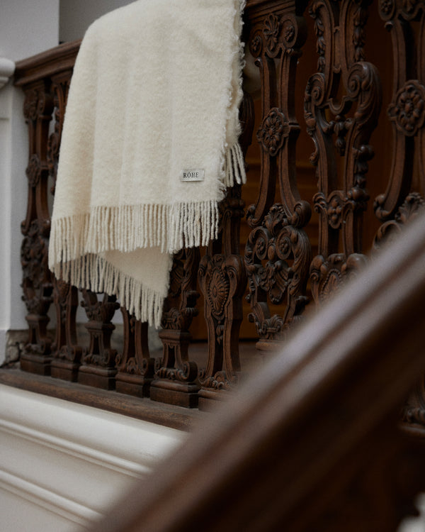 Cream wool blanket with chunky fringes, woven label RÔHE, draped over ornately carved dark-wood railing. Made from Italian virgin wool blend.