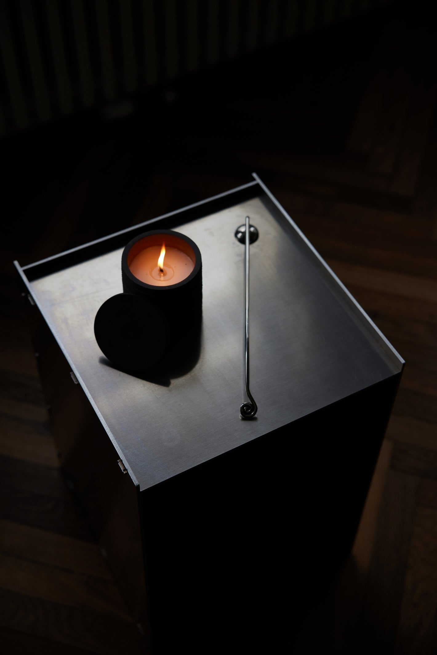 Candle Stopper | silver features an organically curved snuffer with an upward-spiraling handle, placed beside a lit candle on a brushed-steel surface.
