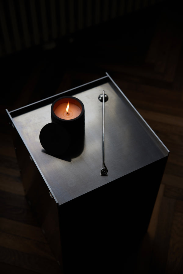 Candle Stopper | silver features an organically curved snuffer with an upward-spiraling handle, placed beside a lit candle on a brushed-steel surface.