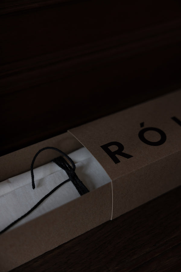 Candle Stopper | silver in kraft-paper box, partially opened to reveal a tissue-wrapped object, tied with black cord. Ideal for extinguishing Róhe Scented Candle.