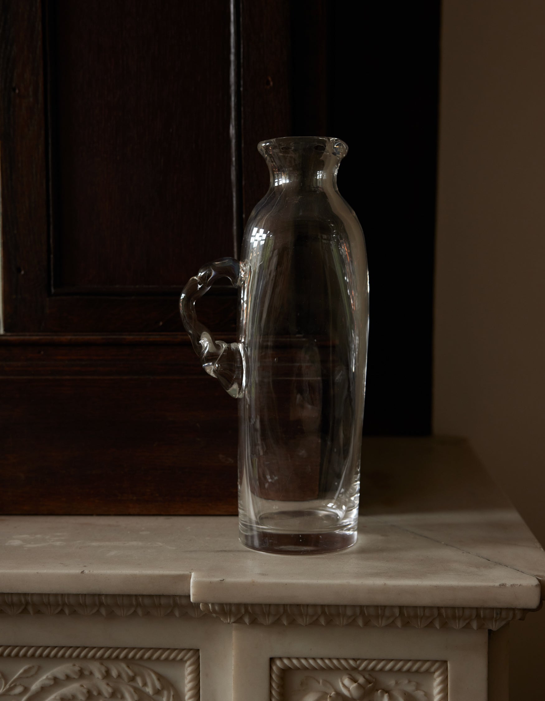 Large water carafe | Glass, a tall, hand-blown pitcher with a curving handle, sits empty on a decorative mantel, contrasting against a dark wooden background.