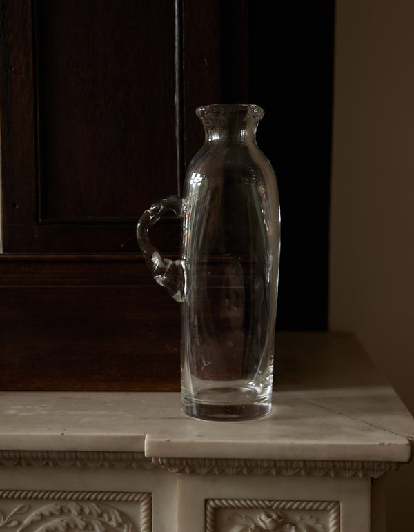 Large water carafe | Glass, a tall, hand-blown pitcher with a curving handle, sits empty on a decorative mantel, contrasting against a dark wooden background.