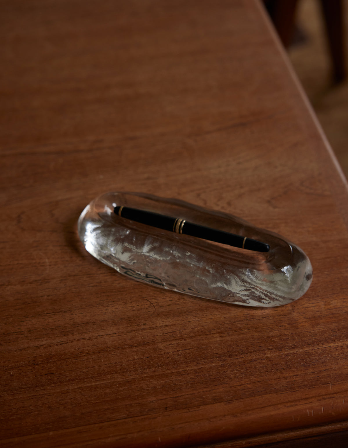 Long trinket | Glass: A sleek clear glass pen rest with an oval indentation, cradling a black fountain pen accented by gold bands, on a wooden tabletop.