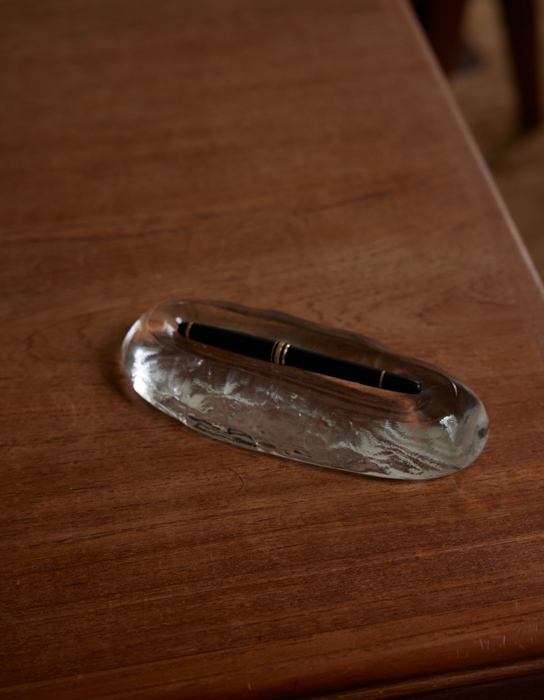 Long trinket | Glass: A sleek clear glass pen rest with an oval indentation, cradling a black fountain pen accented by gold bands, on a wooden tabletop.