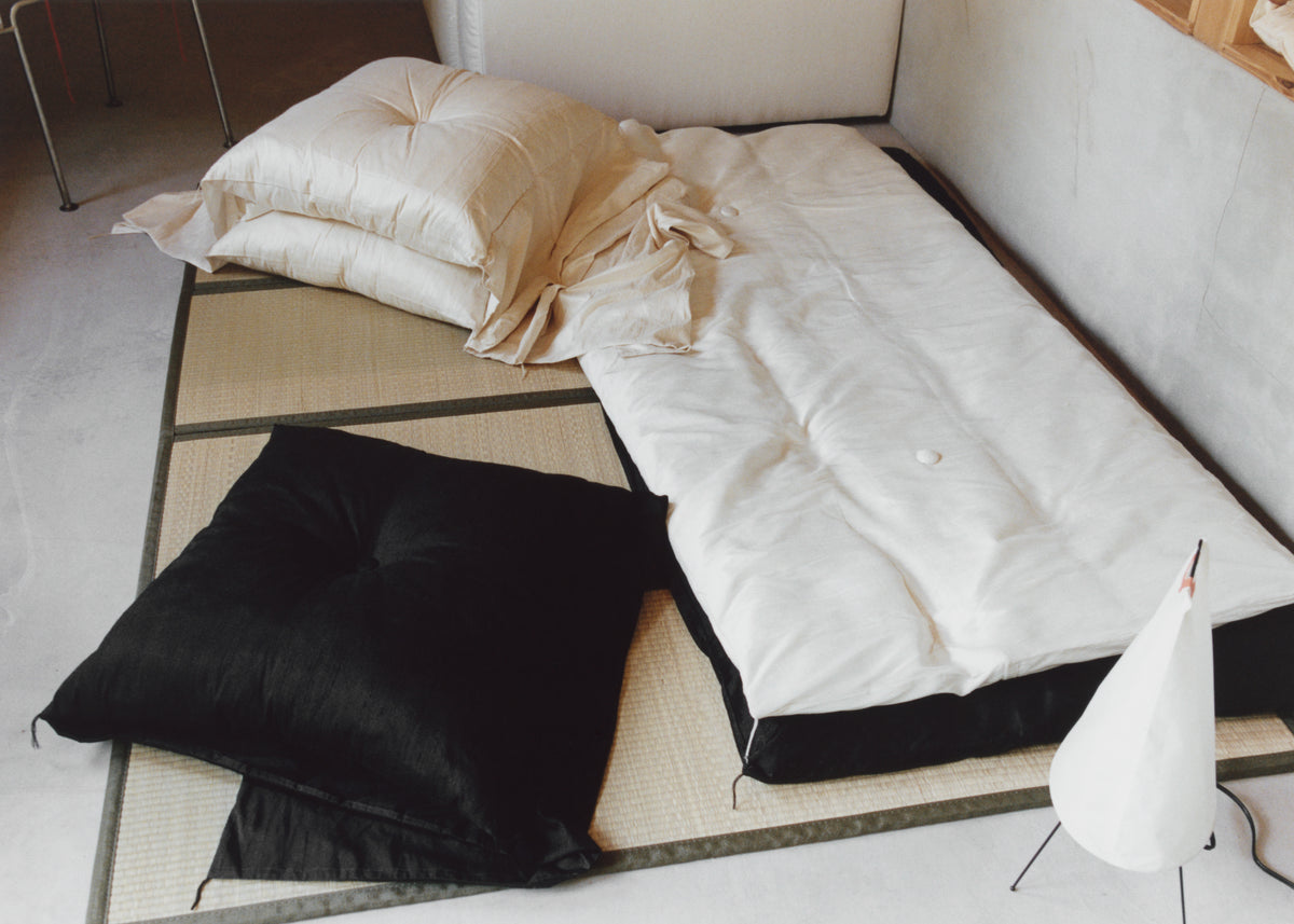 White bedding and pillows arranged on a floor mattress with a black cushion and small white flag in a minimalist room