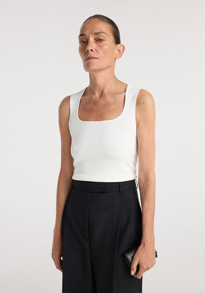 Woman wearing a bustier shaped knitted top | optic white paired with black tailored pants, standing against a plain light background with a neutral expression.