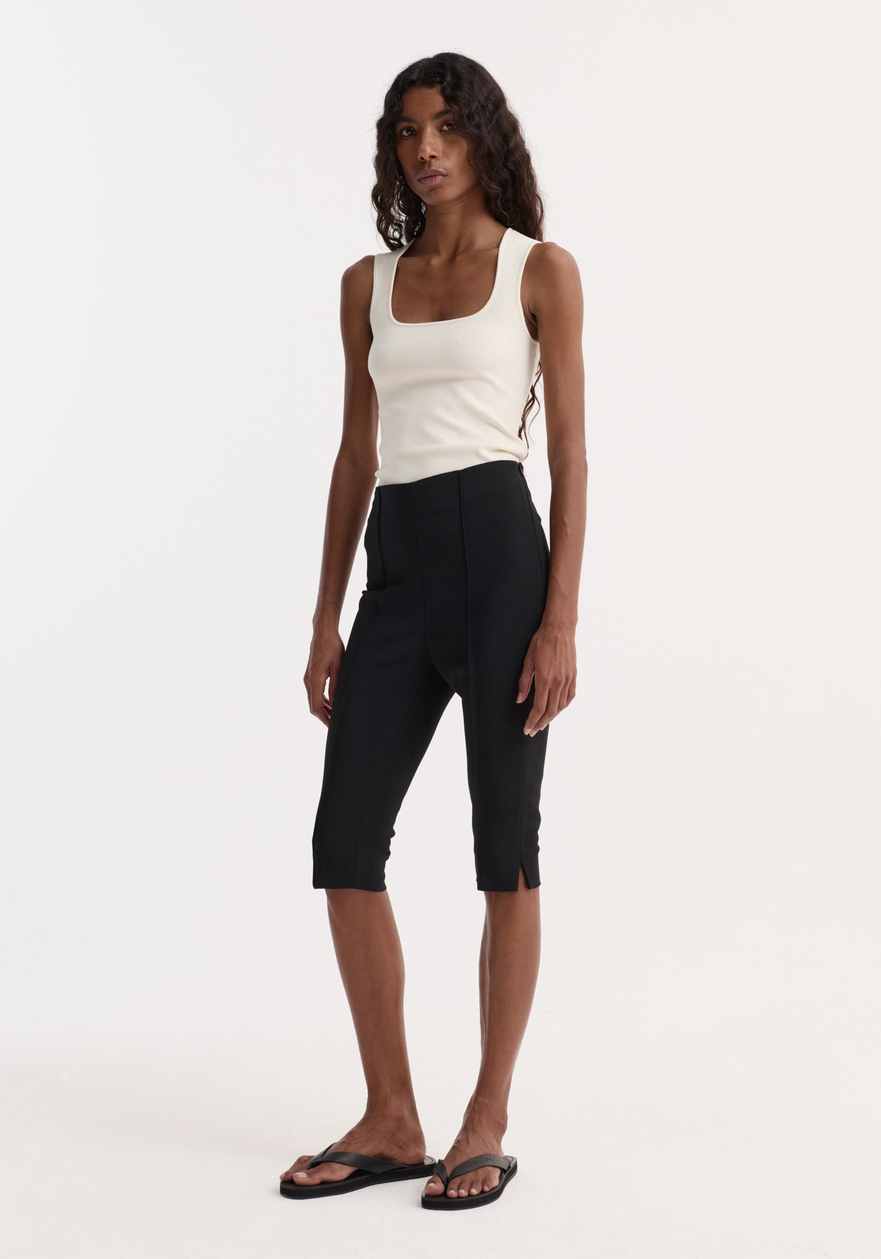 Woman wearing a sleeveless bustier shaped knitted top | off white paired with black cropped pants and black flip-flop sandals standing against a plain background.
