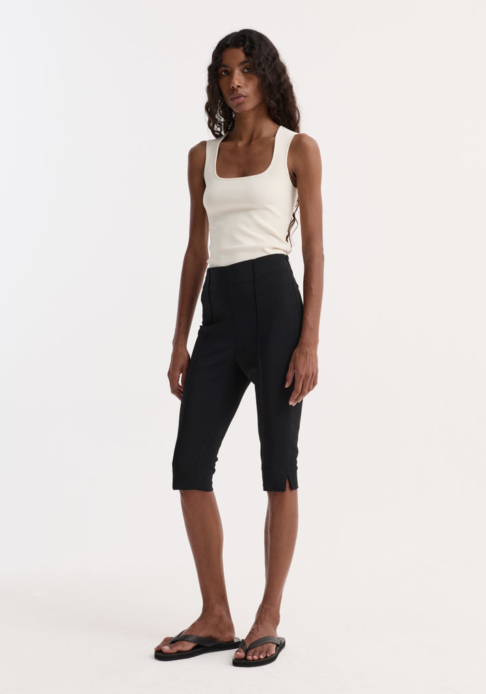 Woman wearing a sleeveless bustier shaped knitted top | off white paired with black cropped pants and black flip-flop sandals standing against a plain background.