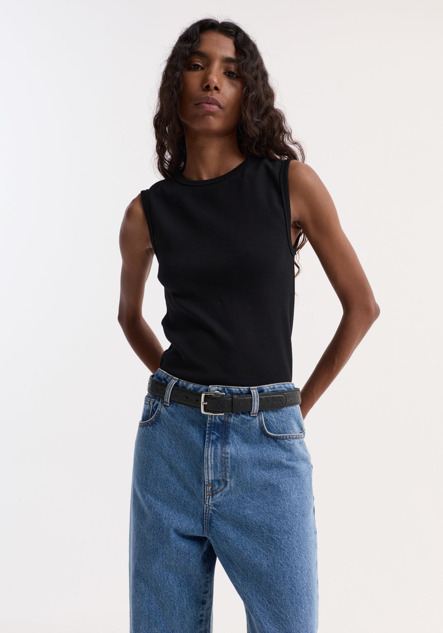 Person wearing Dense rib tank top in black, styled with high-waisted blue jeans and a belt, showcasing its fitted, ribbed texture and versatile, comfortable design.