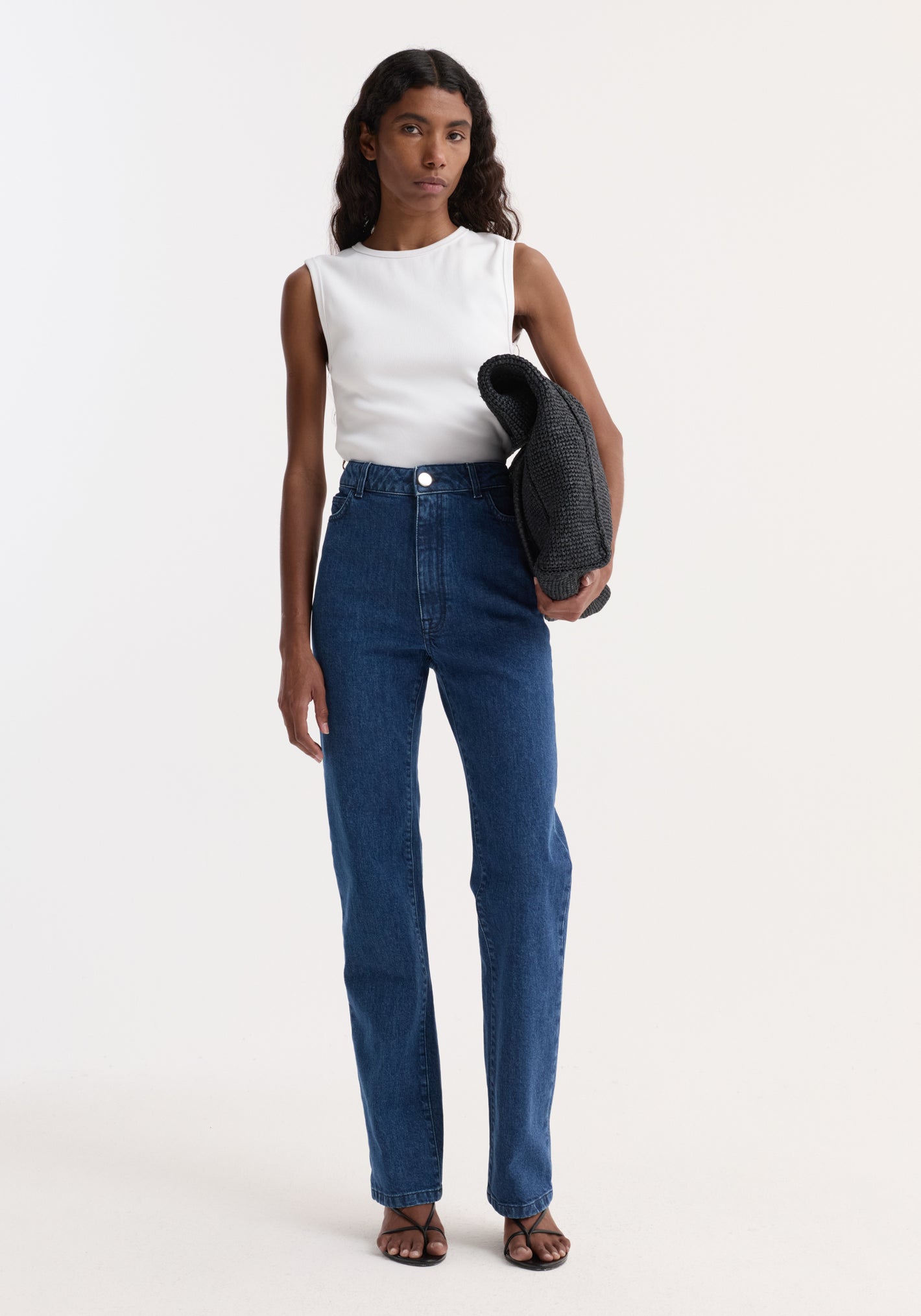 Model wearing Dense rib tank top in optic white, tucked into high-waisted blue jeans, holding a folded dark-grey knitted garment, showcasing a versatile everyday style.