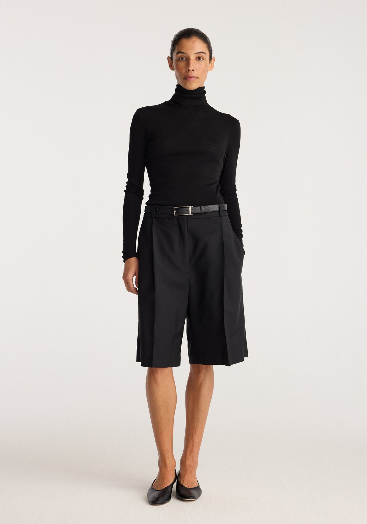 Fine merino turtleneck | black worn by a model with tailored black shorts and black shoes against a neutral studio background