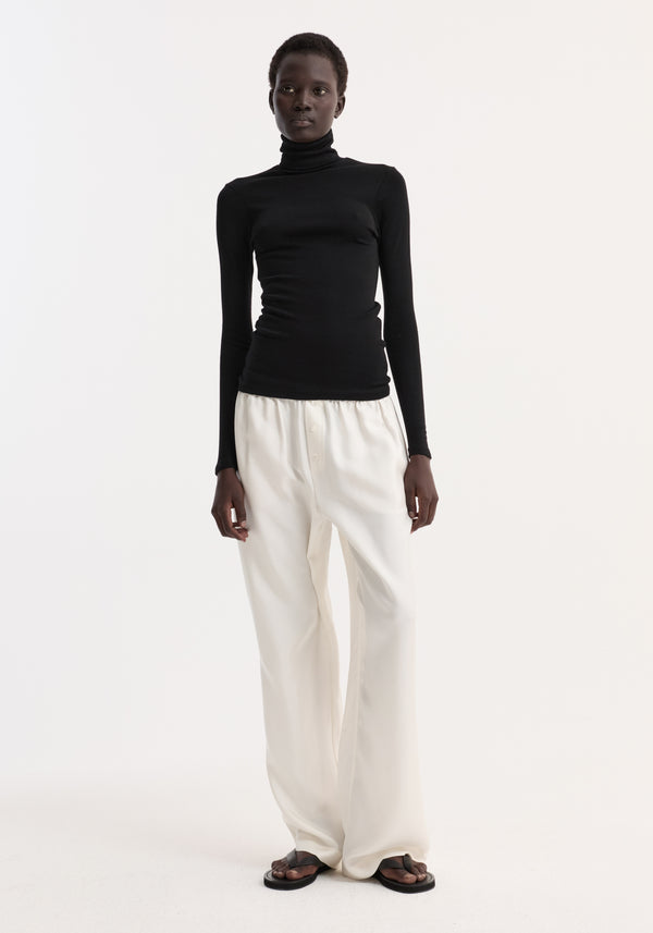 Fine merino turtleneck in black worn by a model, showcasing its fitted long-sleeve design with high-waisted white trousers and black sandals.