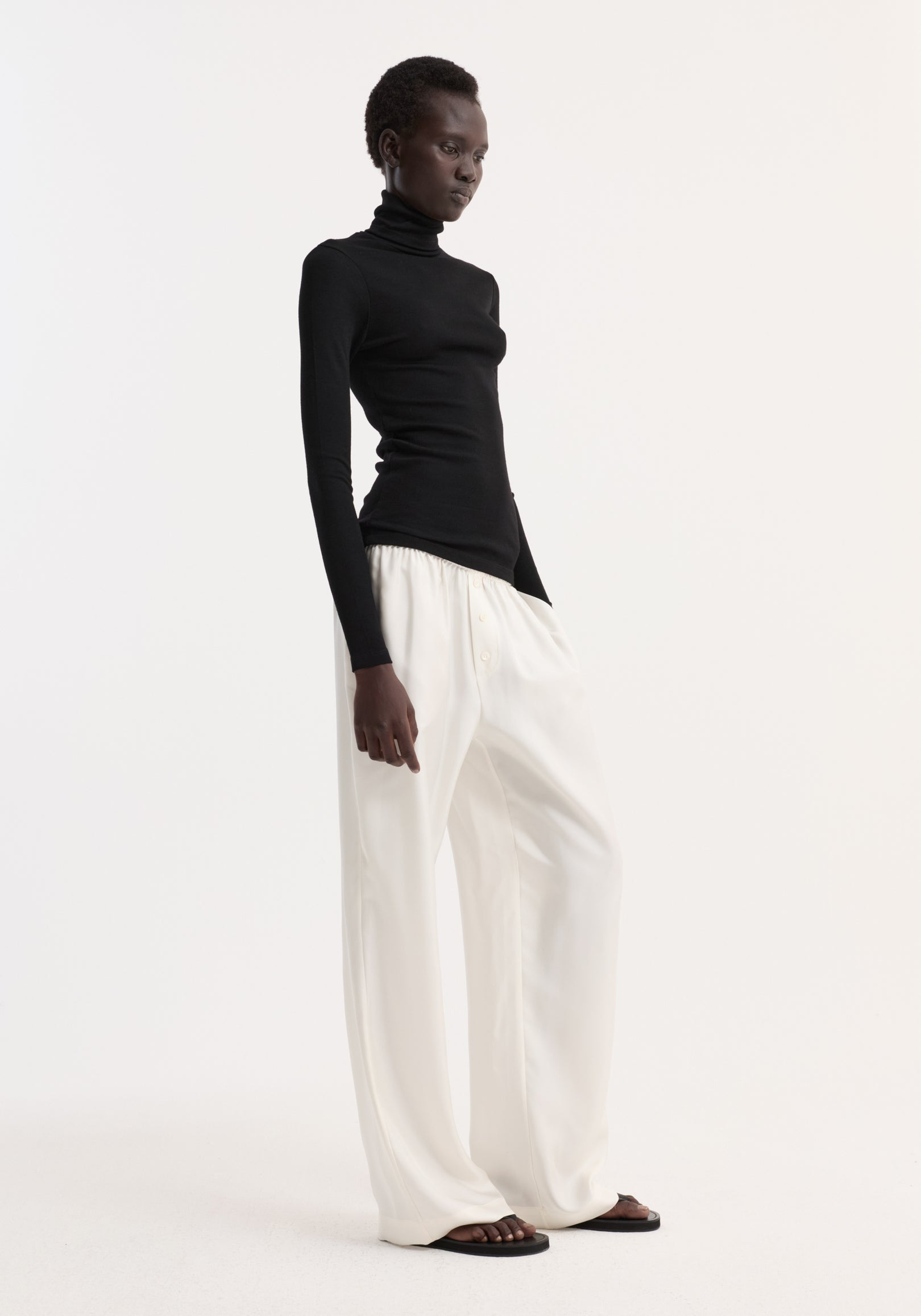 Model wearing a fitted black Fine merino turtleneck | black paired with loose white pants and black sandals against a plain white background.