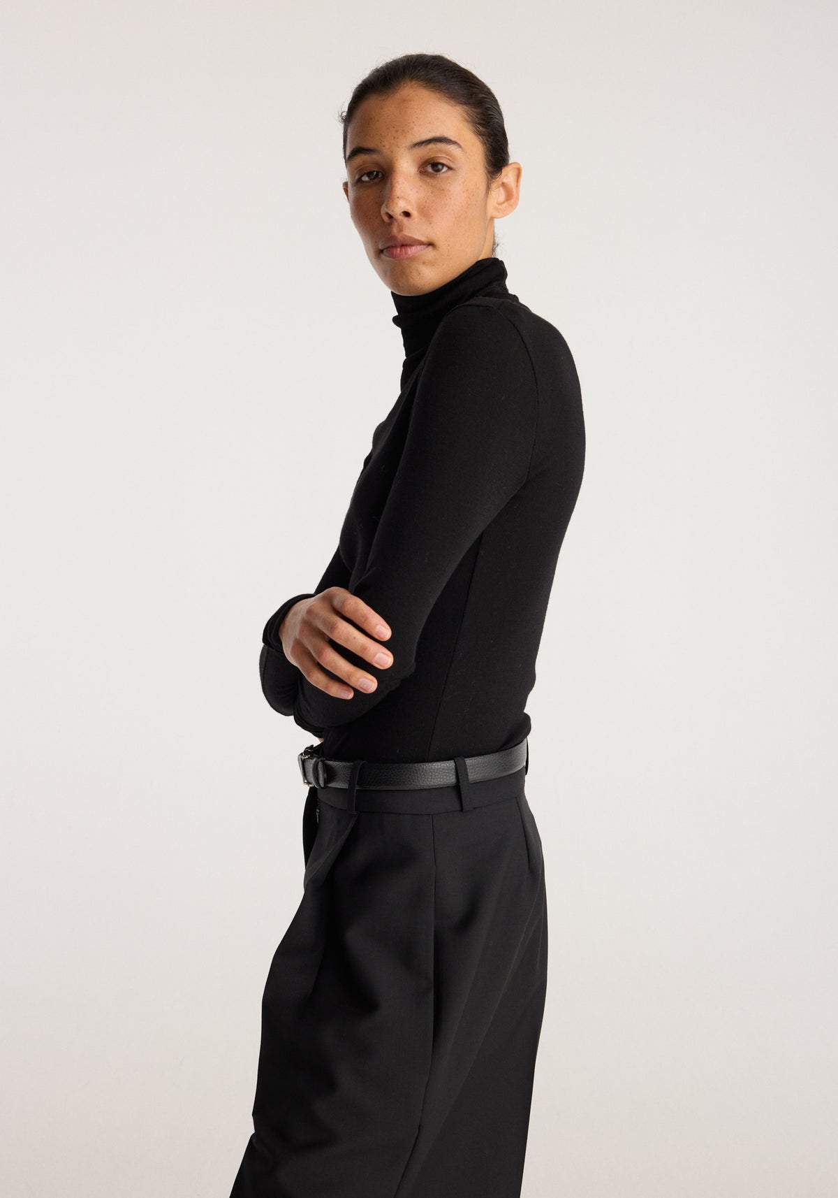 Fine merino turtleneck | black worn by a model in a side profile with arms crossed, styled with black trousers and a belt against a light background