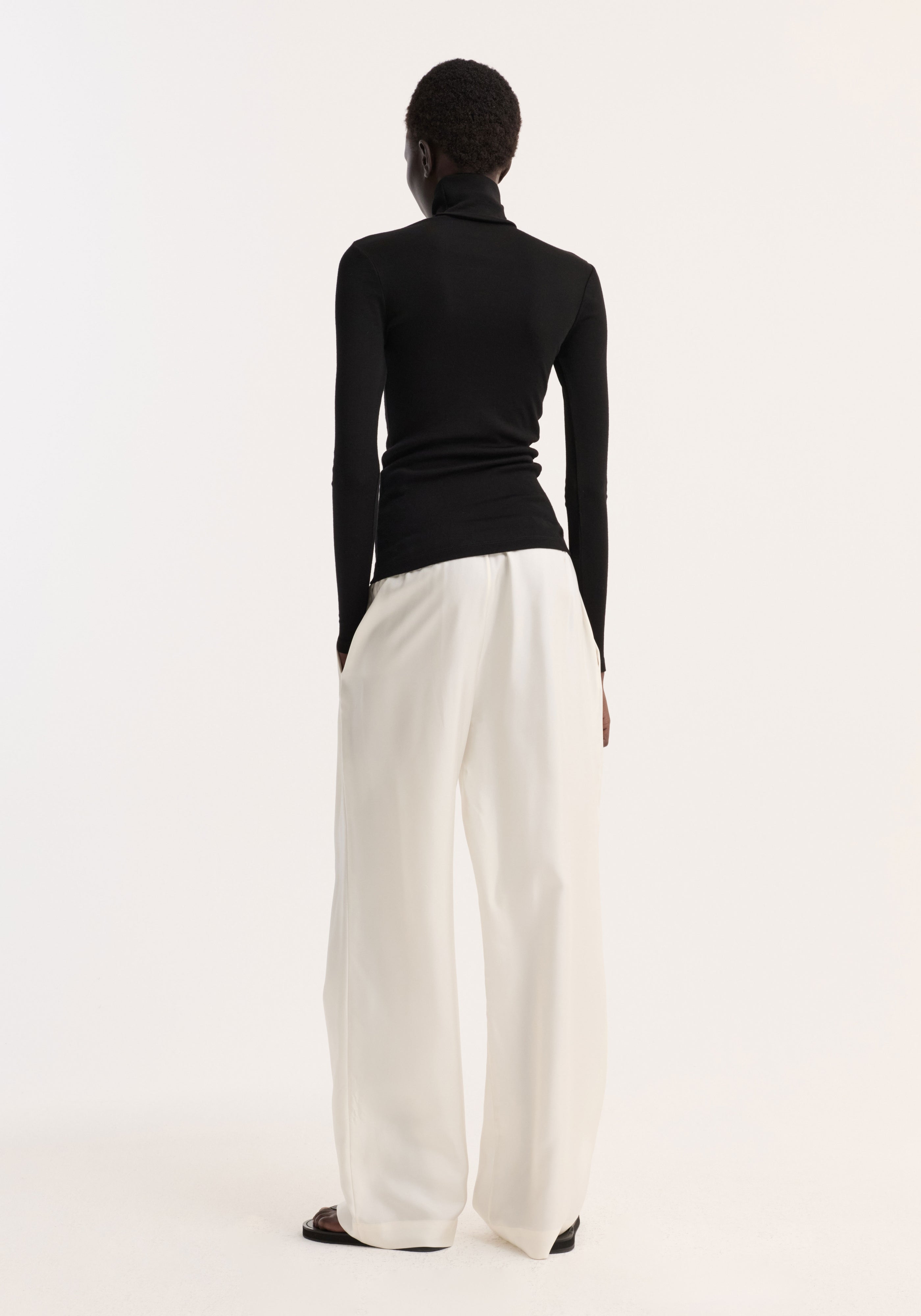 Rear view of a person wearing a fitted black Fine merino turtleneck | black paired with loose white wide-leg pants against a plain background.