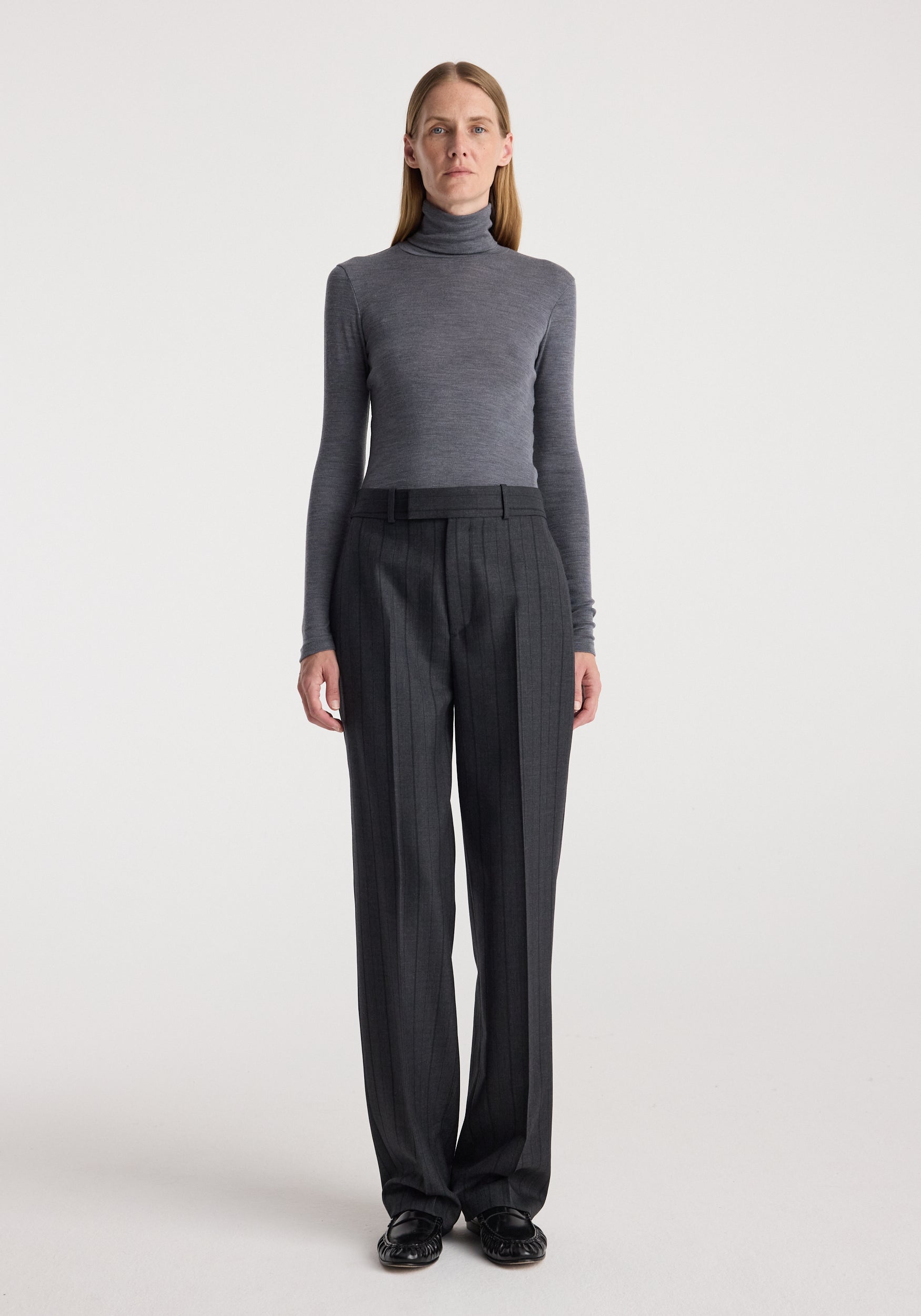 Woman wearing a fitted Fine merino turtleneck | mid grey melange with long sleeves, paired with dark high-waisted trousers, standing against a plain light background.