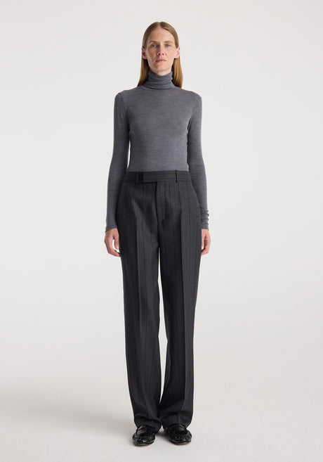 Woman wearing a fitted Fine merino turtleneck | mid grey melange with long sleeves, paired with dark high-waisted trousers, standing against a plain light background.