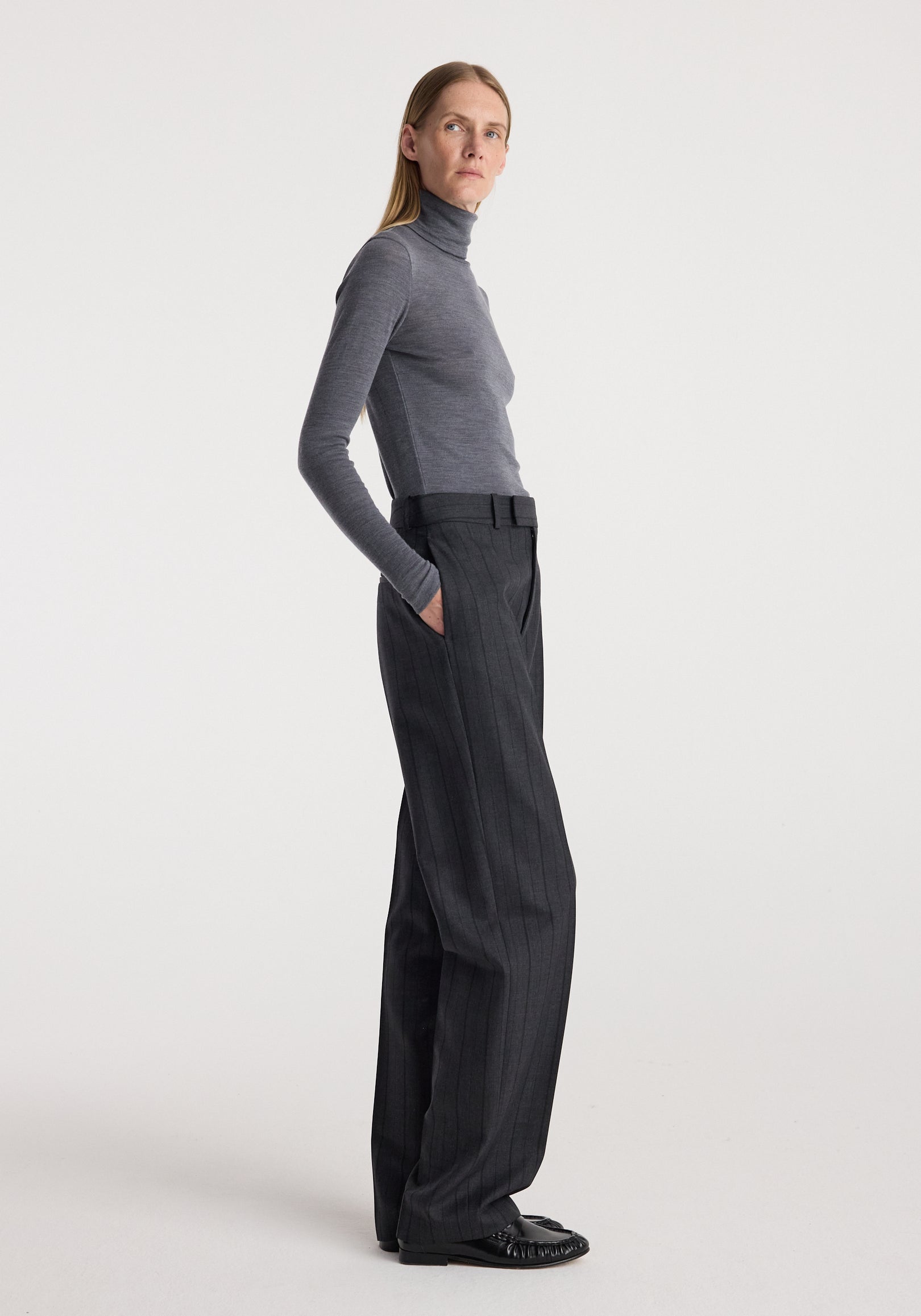 Woman wearing a slim fit Fine merino turtleneck | mid grey melange paired with high-waisted dark trousers and black shoes, standing in a neutral studio setting.