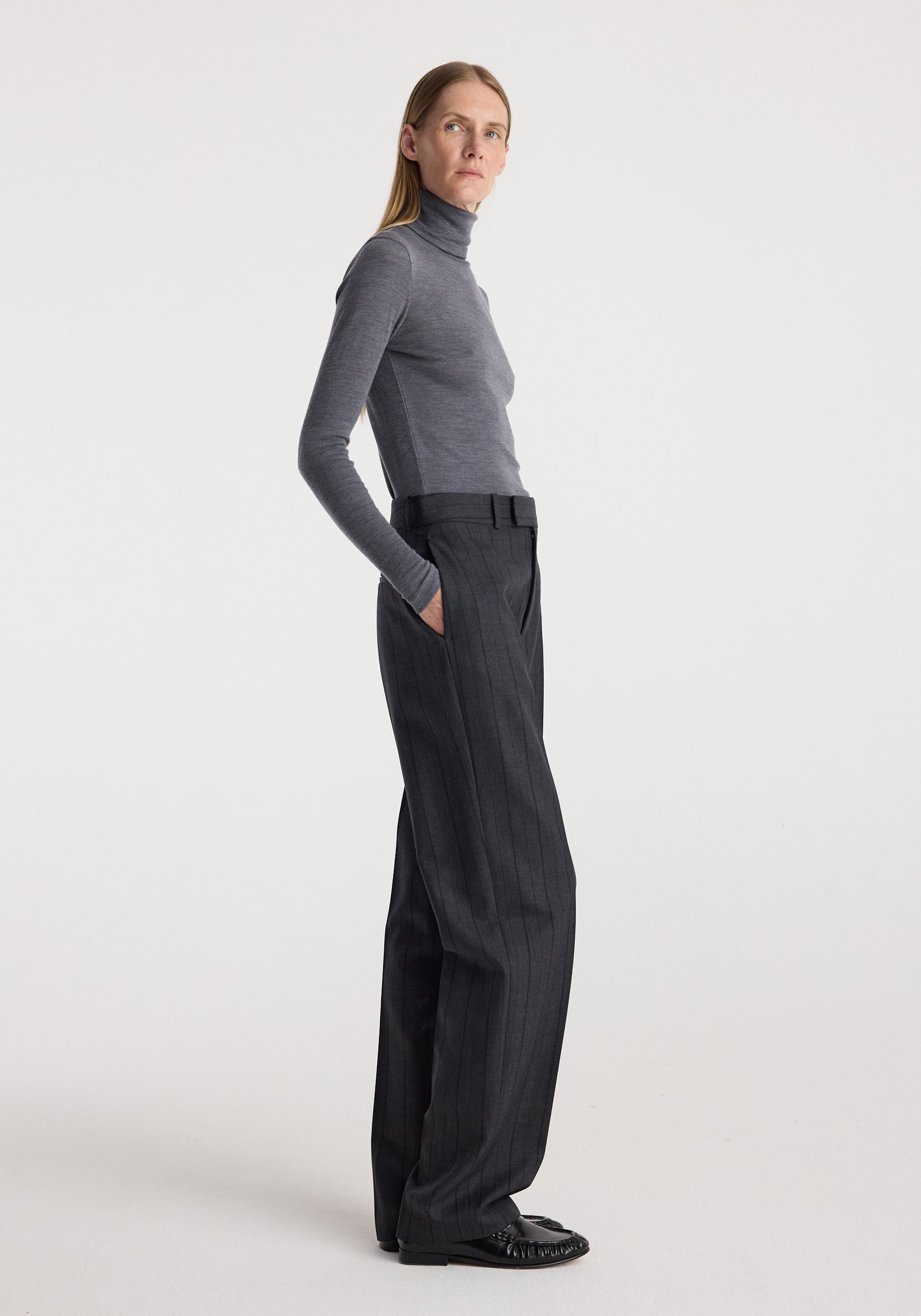 Woman wearing a slim fit Fine merino turtleneck | mid grey melange paired with high-waisted dark trousers and black shoes, standing in a neutral studio setting.