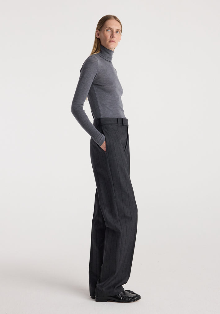 Woman wearing a slim fit Fine merino turtleneck | mid grey melange paired with high-waisted dark trousers and black shoes, standing in a neutral studio setting.