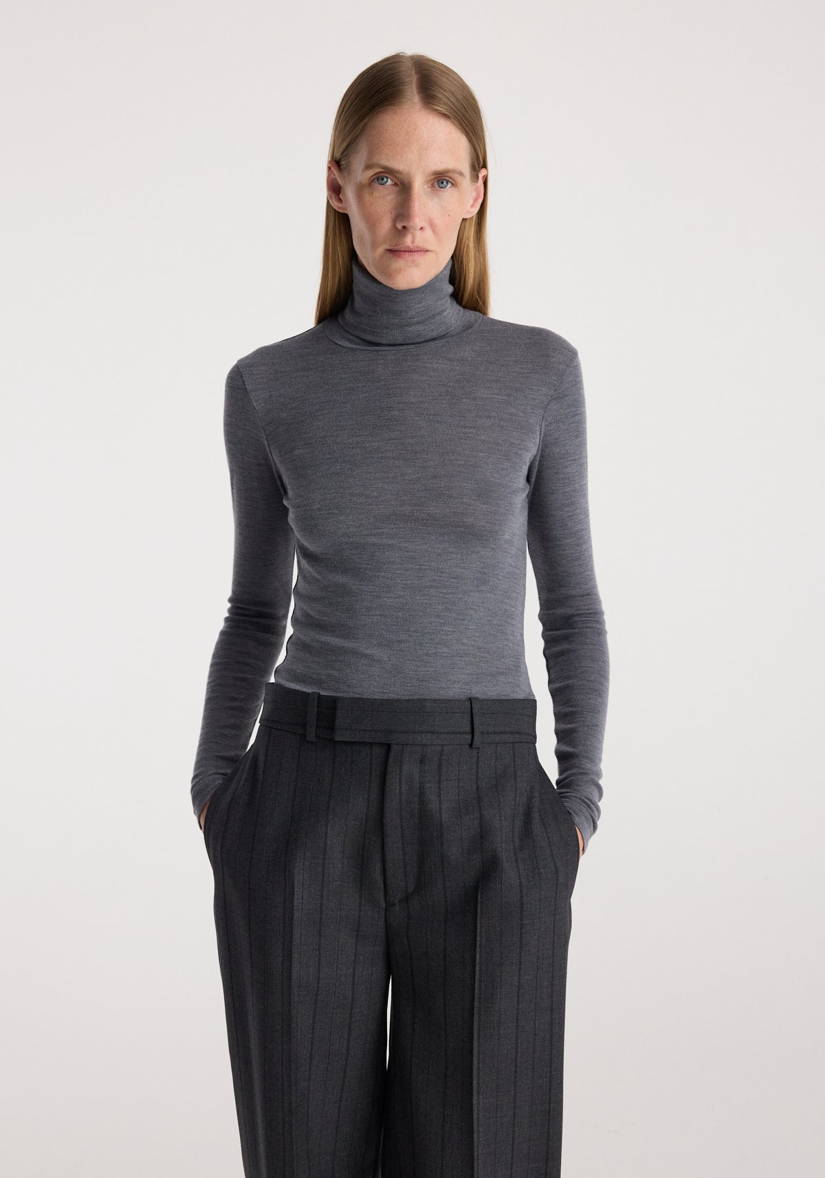 Woman wearing a fitted fine merino turtleneck | mid grey melange paired with high-waisted dark pinstripe trousers against a plain background.