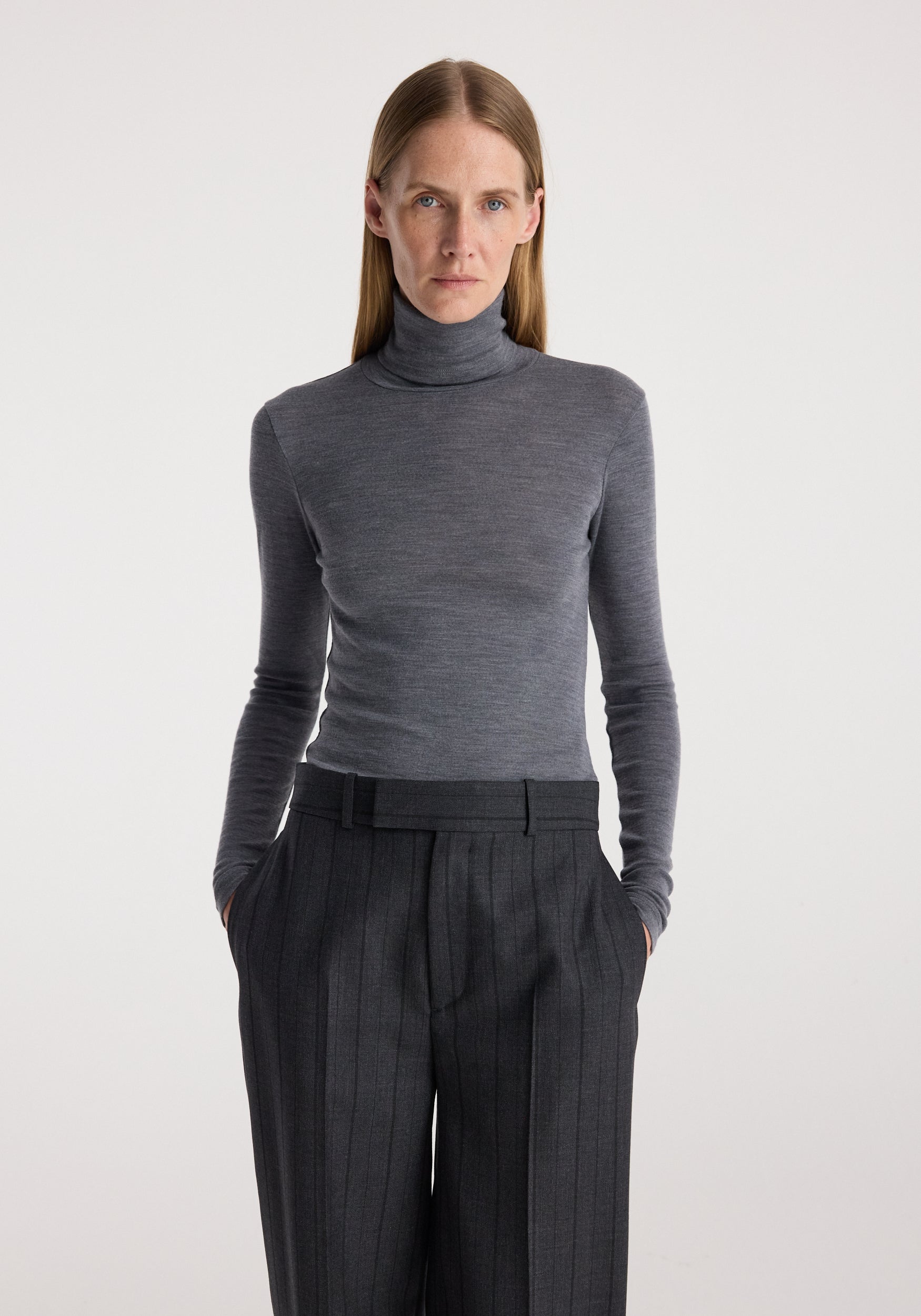 Woman wearing a fitted fine merino turtleneck | mid grey melange paired with high-waisted dark pinstripe trousers against a plain background.