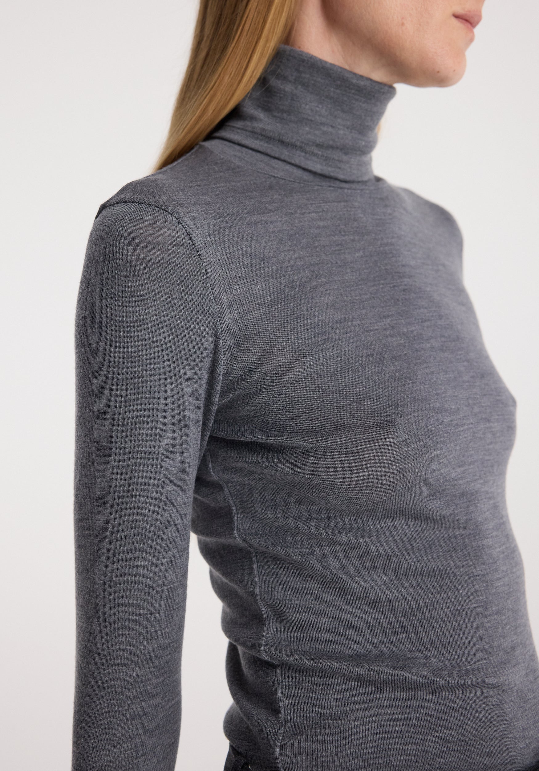 Woman wearing a fitted fine merino turtleneck | mid grey melange with long sleeves and a high collar, shown from the side against a plain background.