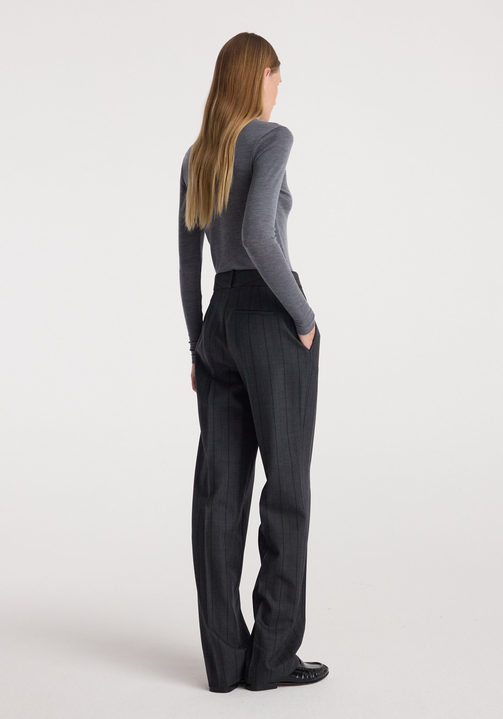 Woman wearing a fine merino turtleneck | mid grey melange, styled with high-waisted dark pinstripe trousers and black shoes, standing with hands in pockets facing away.
