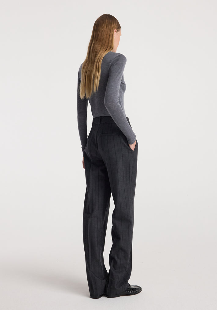 Woman wearing a fine merino turtleneck | mid grey melange, styled with high-waisted dark pinstripe trousers and black shoes, standing with hands in pockets facing away.
