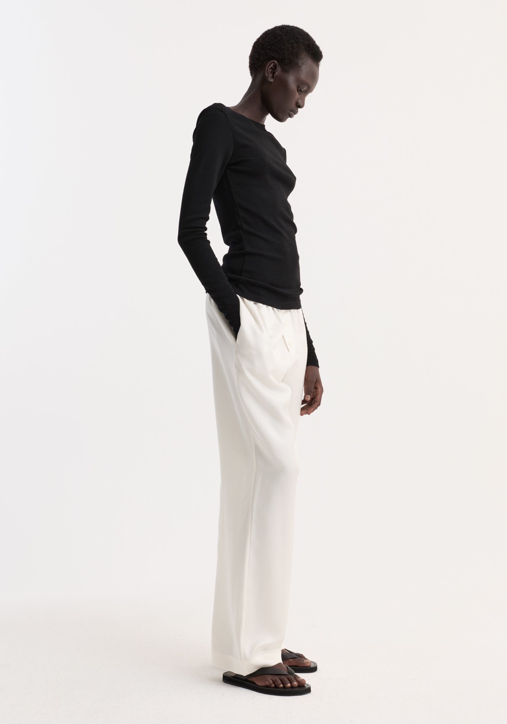 Model wearing a fitted black Fine merino long sleeve | black paired with loose white pants and black sandals against a plain white background.