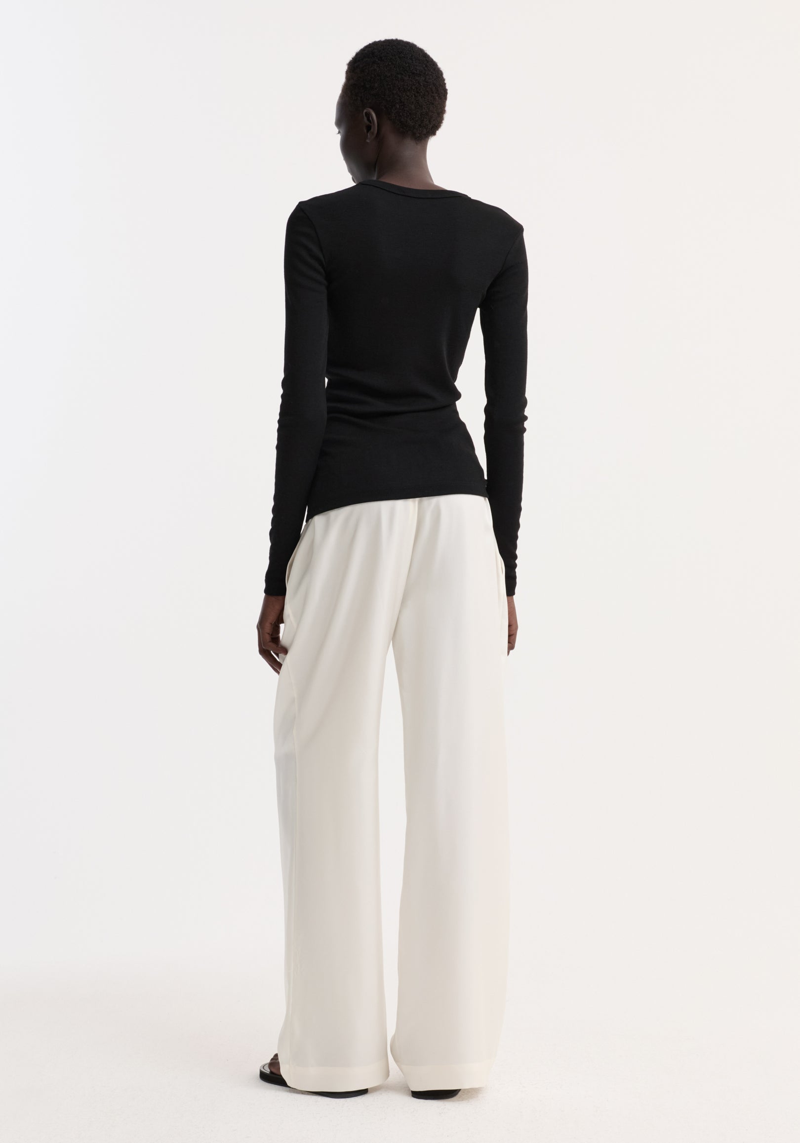 Fine merino long sleeve in black, crew neck, fitted silhouette, worn by model in high-waisted trousers, showcasing minimalist style against a white backdrop.