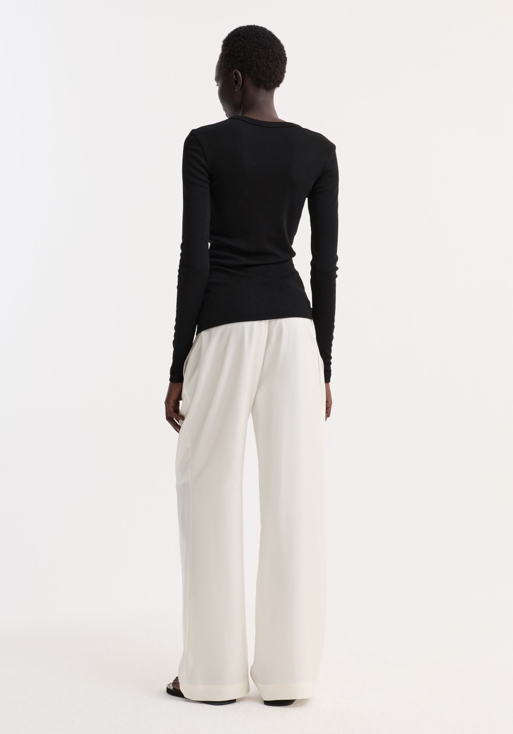 Fine merino long sleeve in black, crew neck, fitted silhouette, worn by model in high-waisted trousers, showcasing minimalist style against a white backdrop.