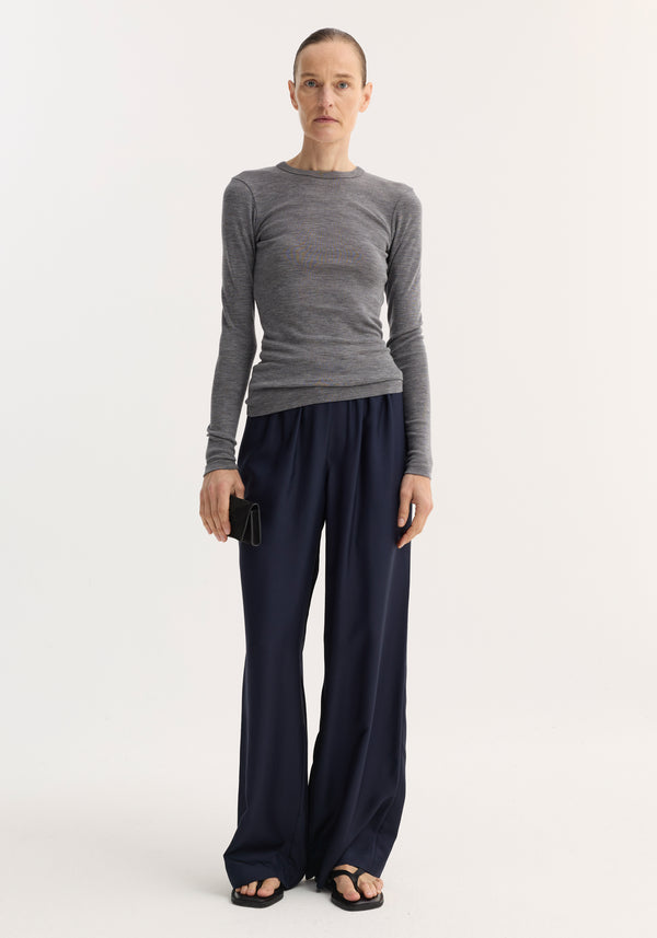 Woman wearing a Fine merino long sleeve in mid grey melange, paired with navy wide-leg trousers, holding a black wallet, looking at the camera.