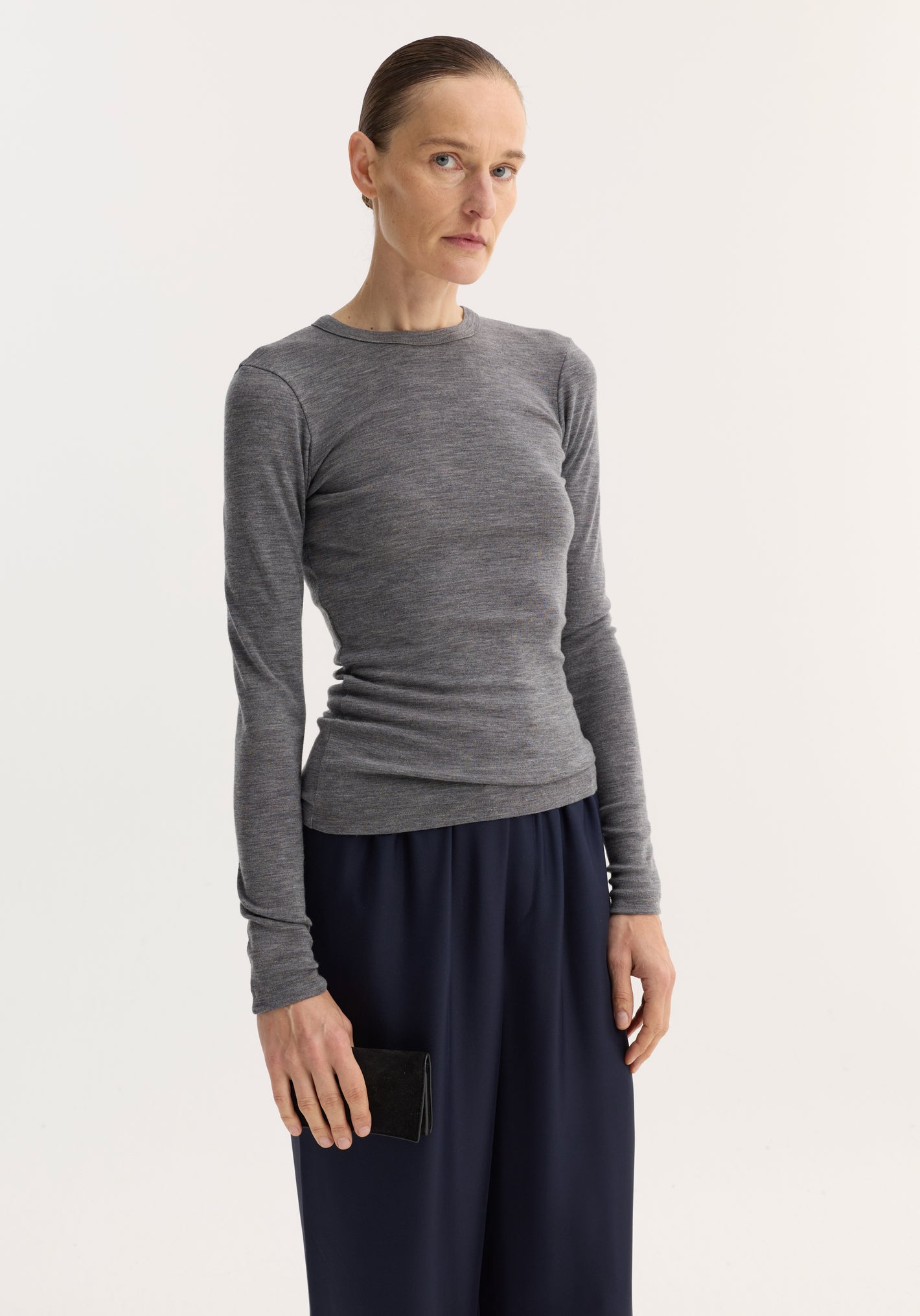 Tall woman in a mid grey melange fine merino long sleeve, showcasing a fitted crew-neck design, paired with wide-leg navy trousers and a black suede clutch.