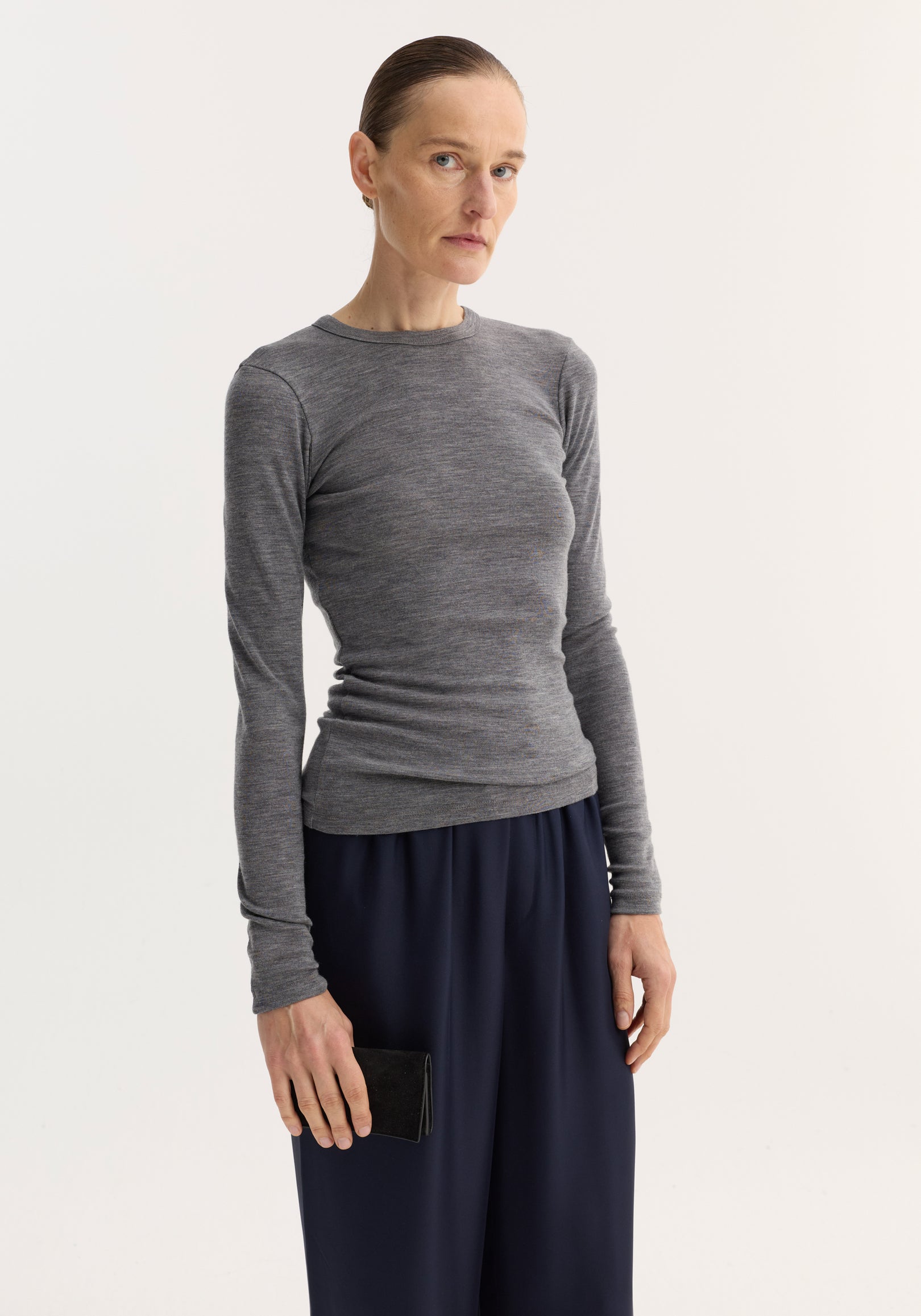 Tall woman in a mid grey melange fine merino long sleeve, showcasing a fitted crew-neck design, paired with wide-leg navy trousers and a black suede clutch.