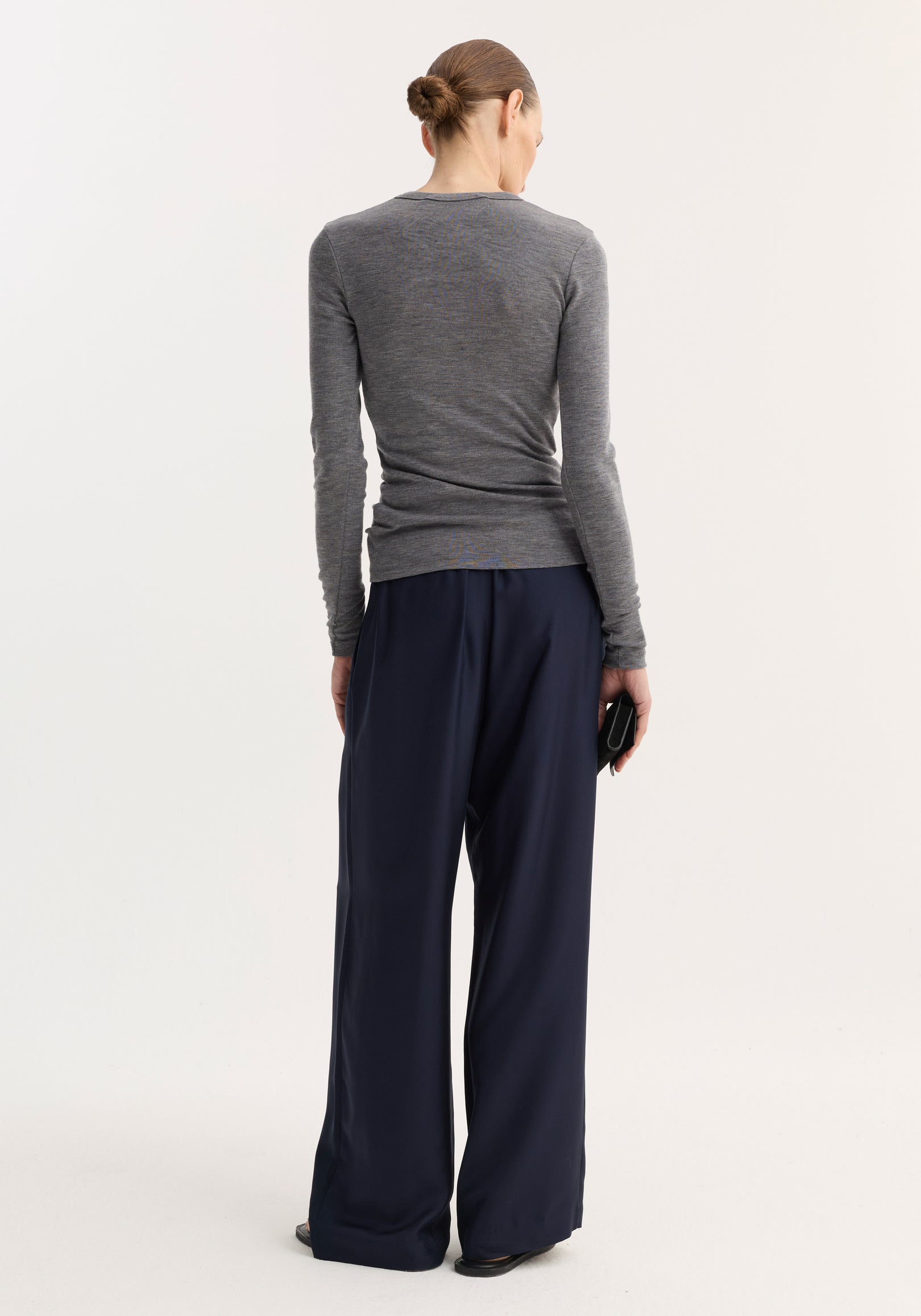 Model in a fitted Fine merino long sleeve in mid grey melange, tucked into wide-leg navy trousers, holding a black clutch, showcasing minimalist style.