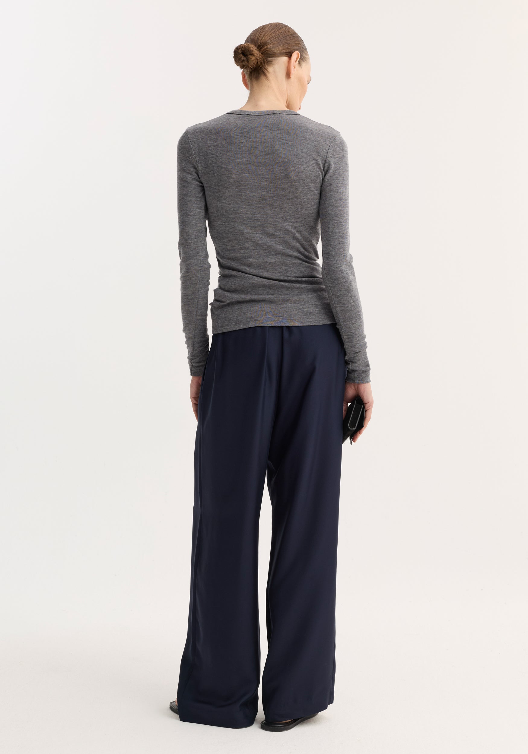 Model in a fitted Fine merino long sleeve in mid grey melange, tucked into wide-leg navy trousers, holding a black clutch, showcasing minimalist style.